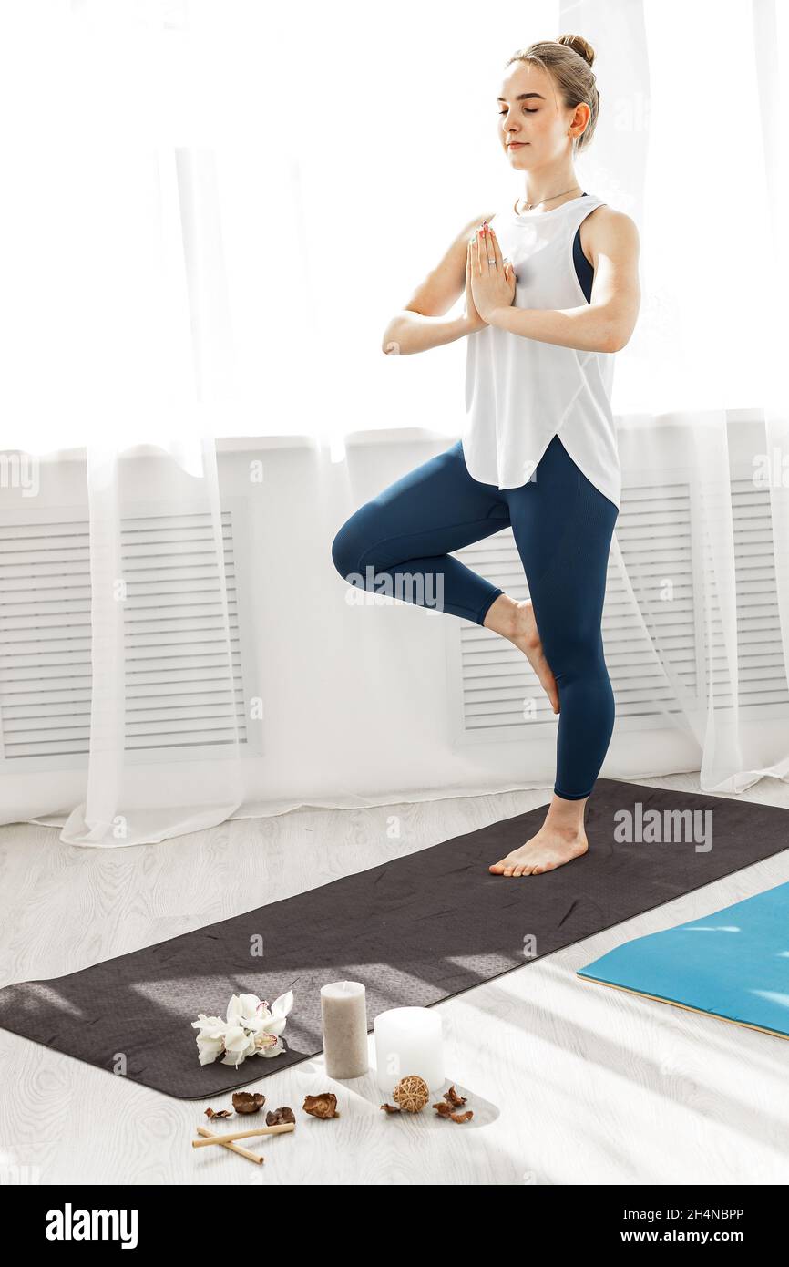 Lächelndes junges sportlich-fittes Mädchen, das Hatha Yoga in einem modernen Yoga-Studio macht. Sie steht in einer Assana auf einem Bein mit geschlossenen Augen. Gesunde Lebensweise Kontra Stockfoto