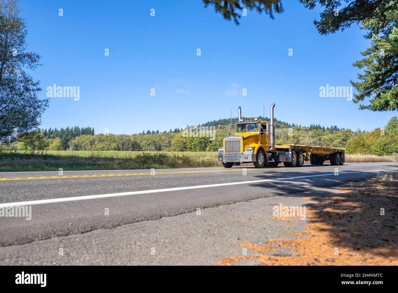 Leistungsstarker gelber Big Rig klassischer Sattelschlepper, der auf der schmalen Straße zum Lager für die nächste Ladung leeres Flachbett-Sattelauflieger transportiert Stockfoto