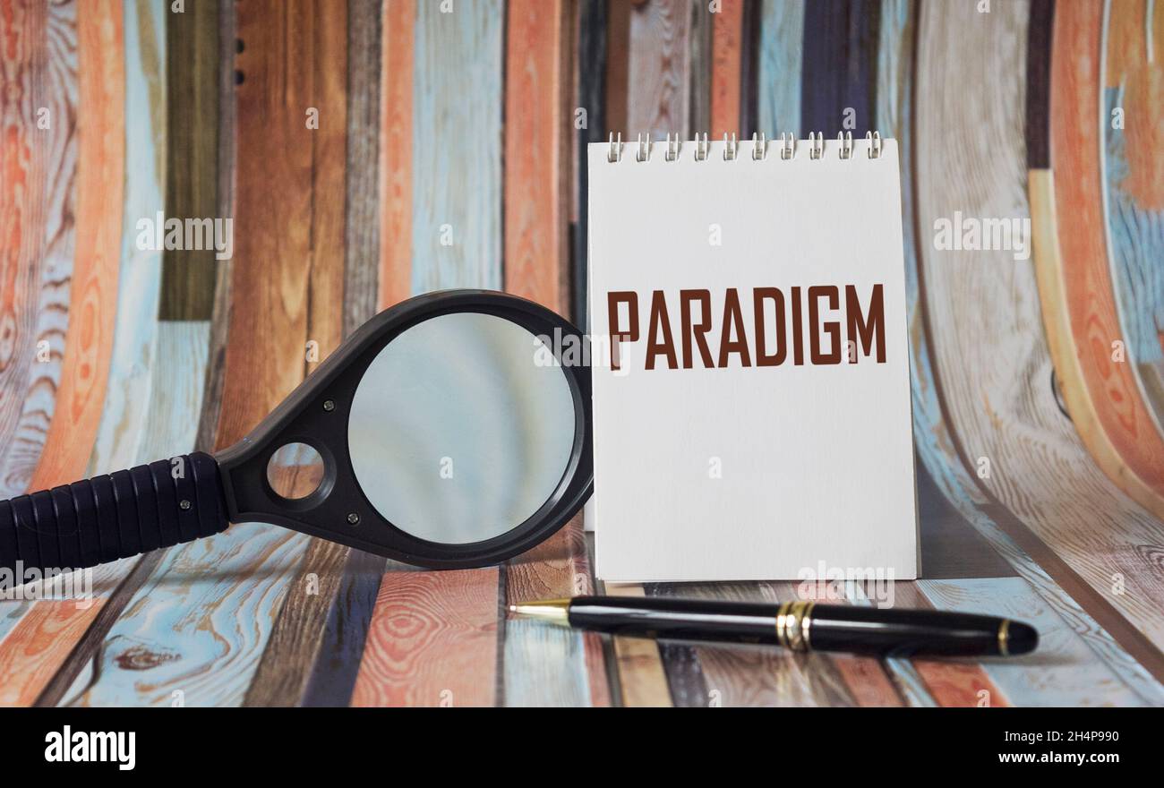 Das Wort Paradigma steht auf einem weißen Notizbuch, neben einem Stift und einer Lupe auf einem gestreiften Hintergrund Stockfoto