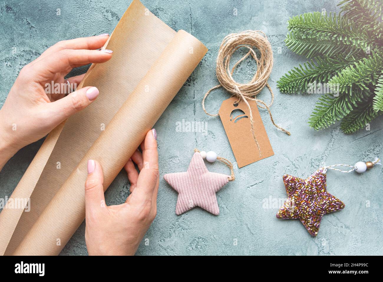 Nahaufnahme der Hände mit einer Rolle Geschenkpapier auf texturiertem Hintergrund mit Weihnachtsspielzeug, Anhänger und Fichtenzweig. Gemütliches Weihnachtskonzept Stockfoto