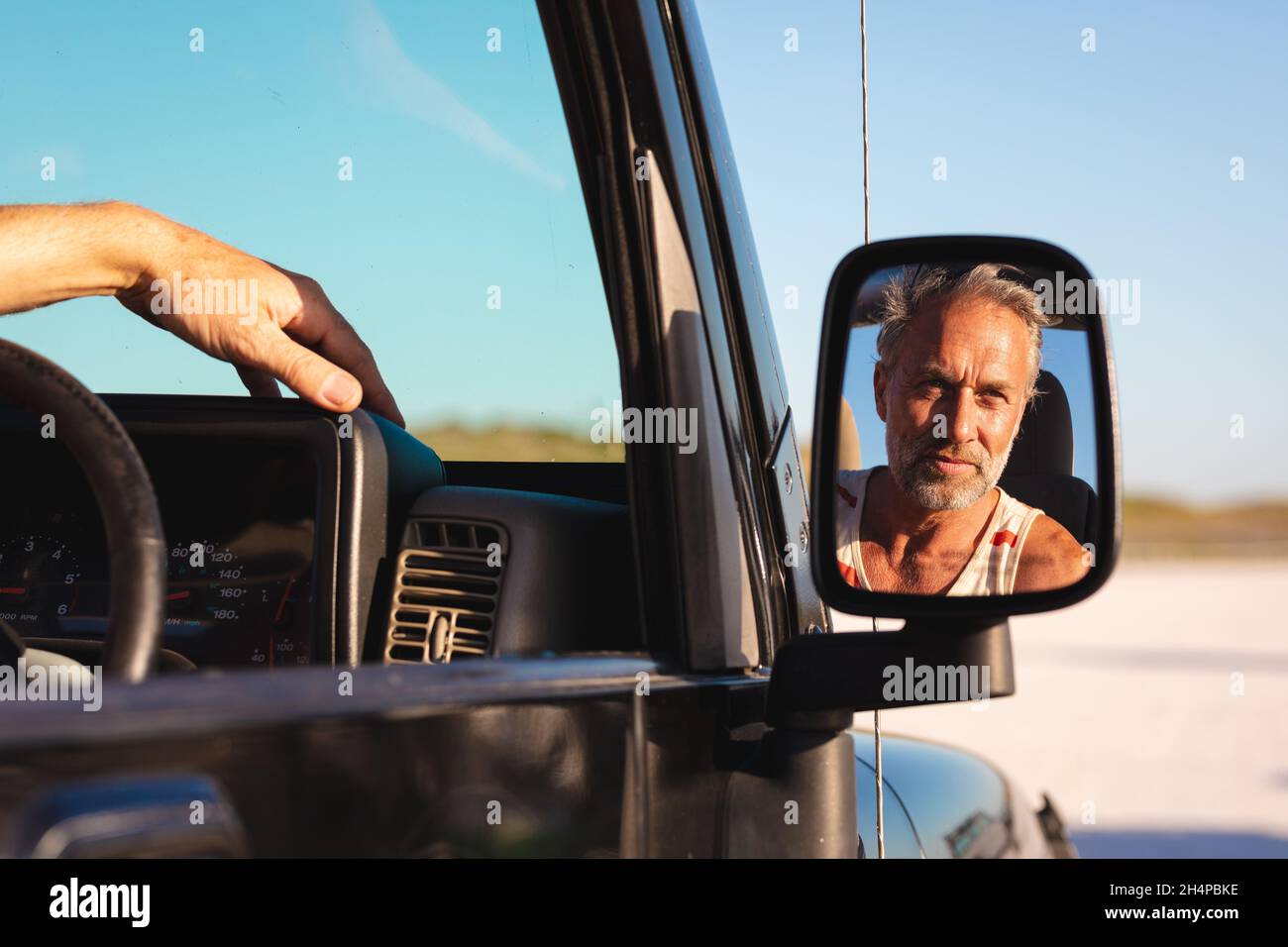 Porträt eines lächelnden kaukasischen Mannes im Auto, der sich am sonnigen Tag am Meer im Seitenspiegel widerspiegelt Stockfoto