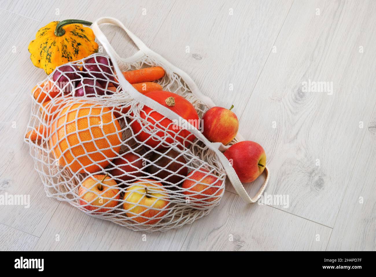 Orangenkürbis, Äpfel, Zwiebeln, Zucchini und Karotten in einem natürlichen wiederverwendbaren Netzbeutel. Hintergrund für gesunde Lebensmittel. Weißer Holzhintergrund. Stockfoto