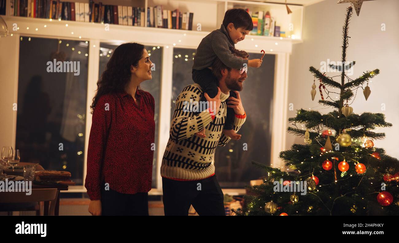 Mann mit seinem Sohn auf den Schultern, der ihm hilft, den Weihnachtsbaum zu schmücken. Innenaufnahme eines Familienhauses, das zu Weihnachten dekoriert wurde. Stockfoto