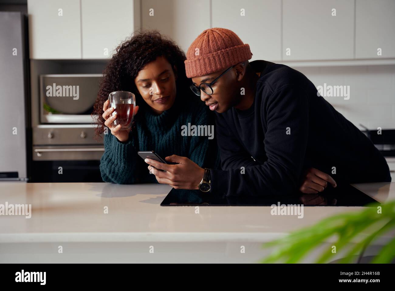 Ein multiethnisches Paar lacht und lächelt am Telefon, während es in der Küche in einer modernen Wohnung ist Stockfoto