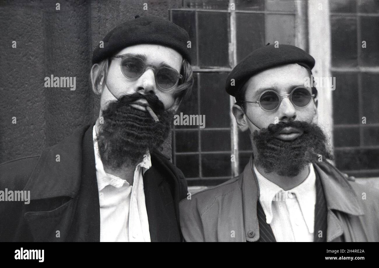 In den 1960er Jahren, historisch, zwei Männer, die vor einem Gebäude in Regenmänteln standen und mit Baretten, Sonnenbrillen und falschen Bärten verkleidet waren, plus rauchenden kleinen Zigarren, Deutschland. Stockfoto