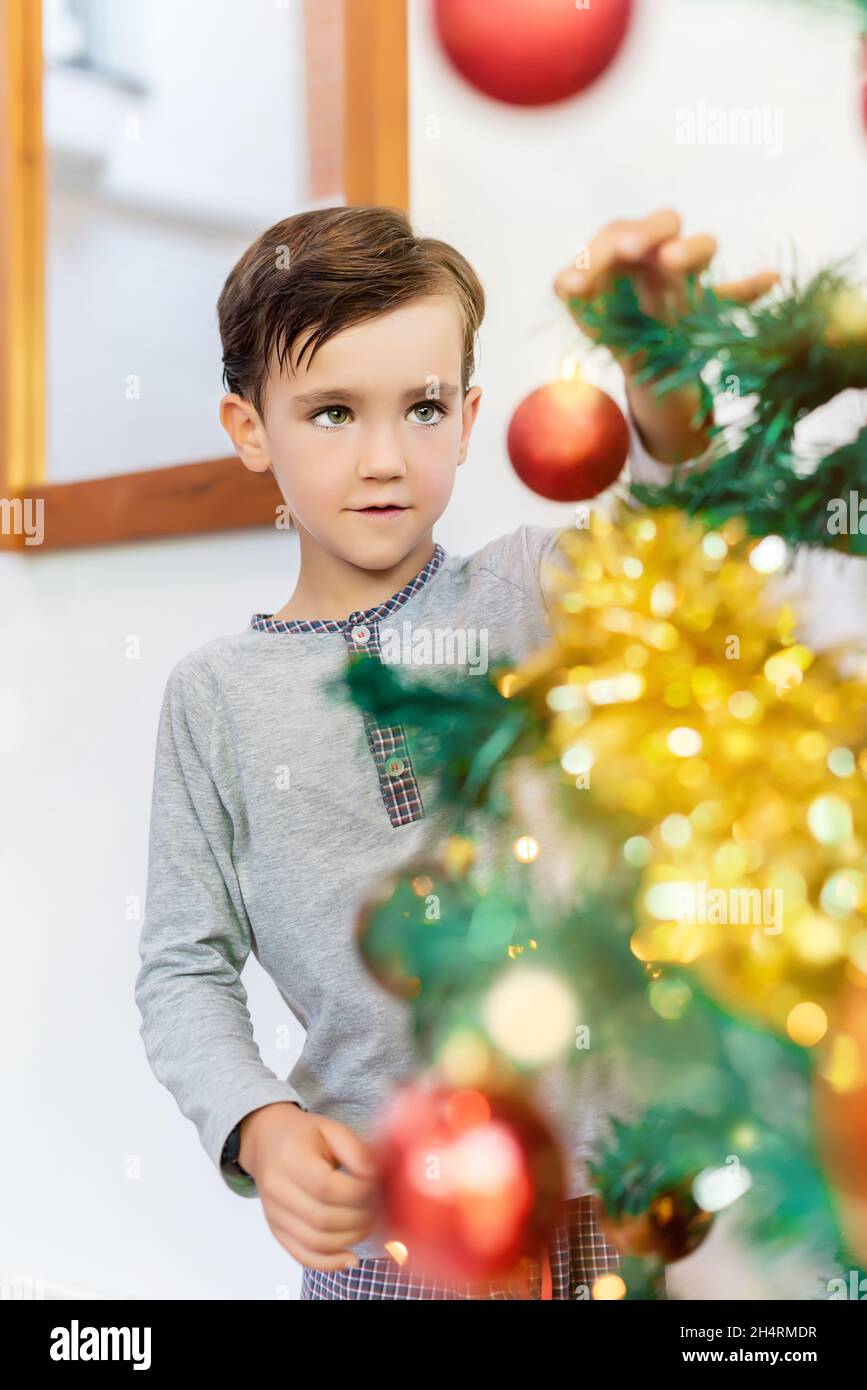 Fröhlicher Junge im Pyjama, der den Weihnachtsbaum schmückt und an einem kalten Wintermorgen Weihnachtsbälle darauf legt Stockfoto