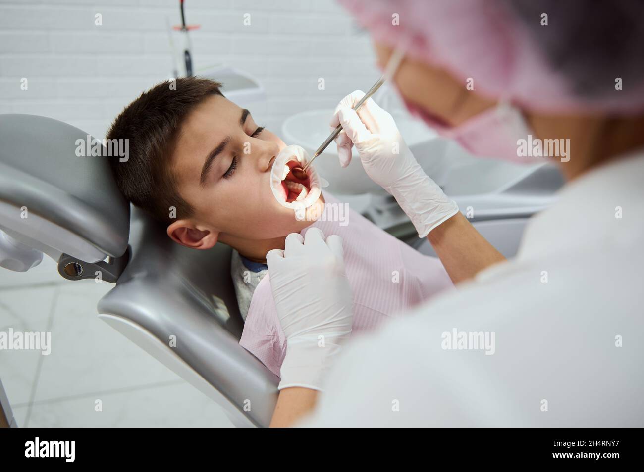 Selbstbewusster Kinderjunge, der auf dem Zahnarztstuhl in der weißen modernen Zahnklinik sitzt, während eine nicht erkennbare weibliche Zahnärztin medizinische Manipulationen und Stockfoto
