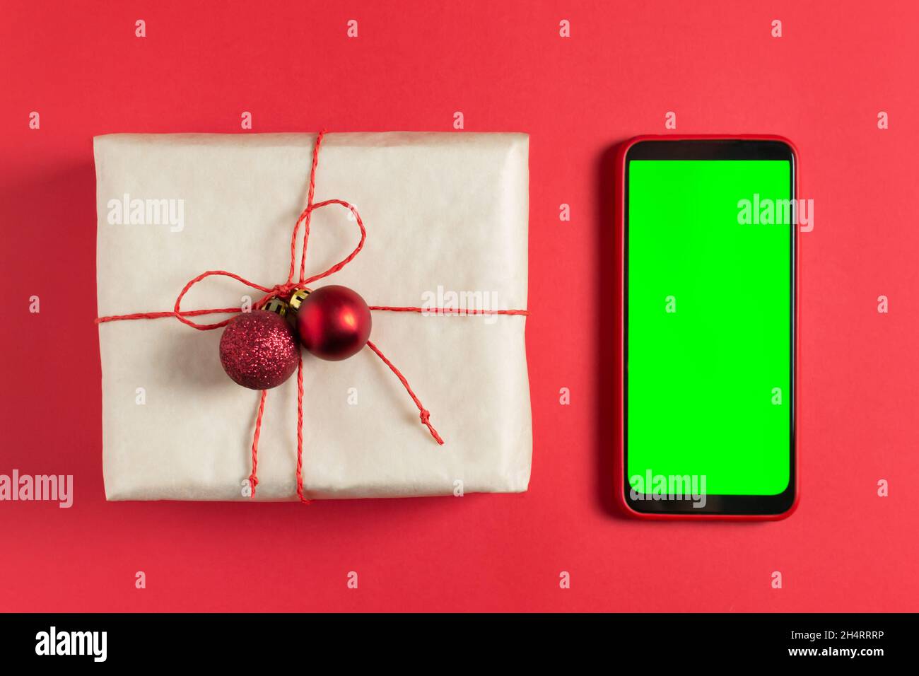 Ein Neujahrsgeschenk ist in Bastelpapier gewickelt und mit rotem Jgut und kleinen Weihnachtskugeln auf rotem Hintergrund gebunden. Handy mit grünem Bildschirm, Chroma Stockfoto