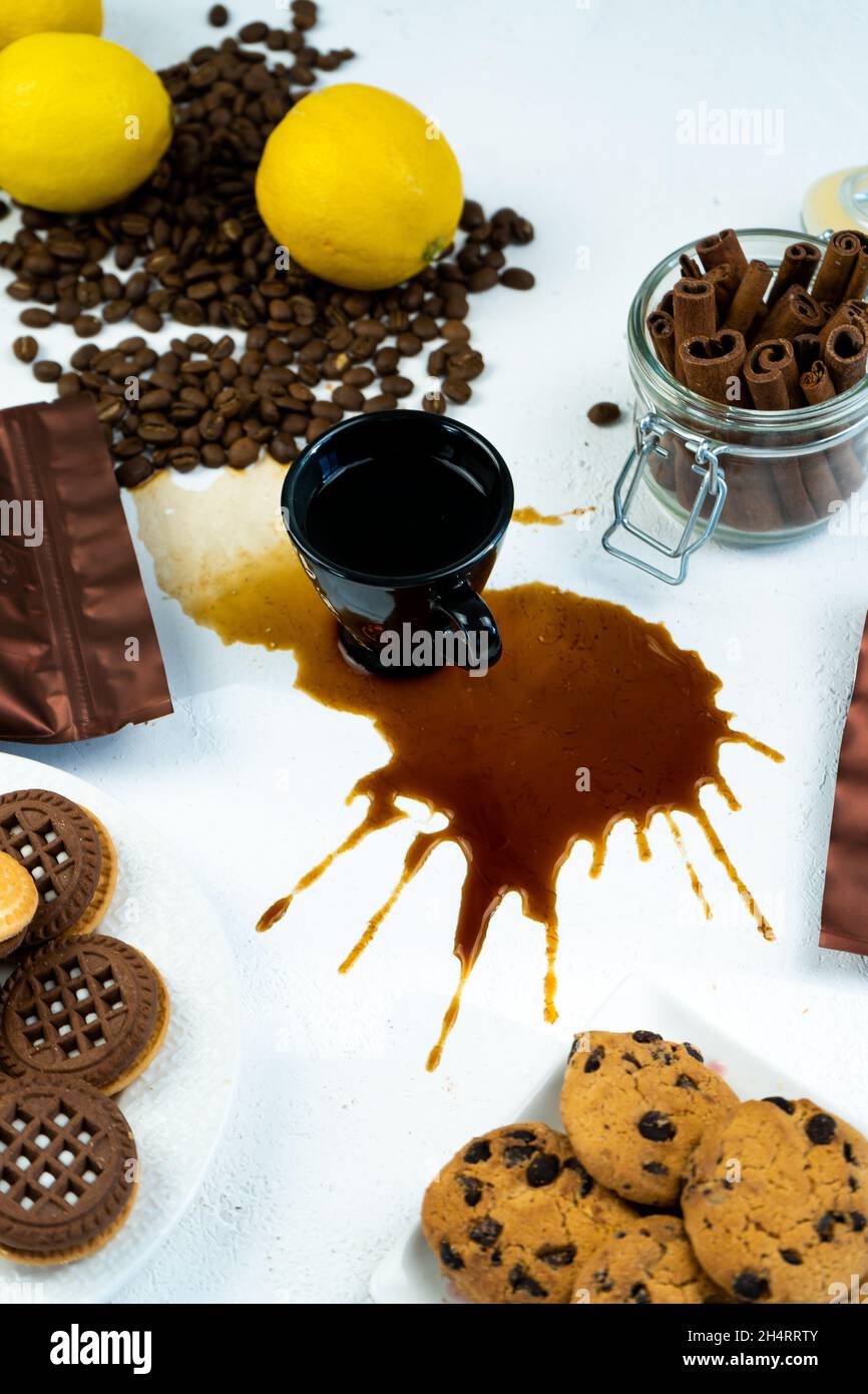 Verschütteter Kaffee aus einer schwarzen Tasse auf einem weißen Tisch. Kaffeebohnen, Zimt und Kekse. Frühstück in der Küche. Stockfoto