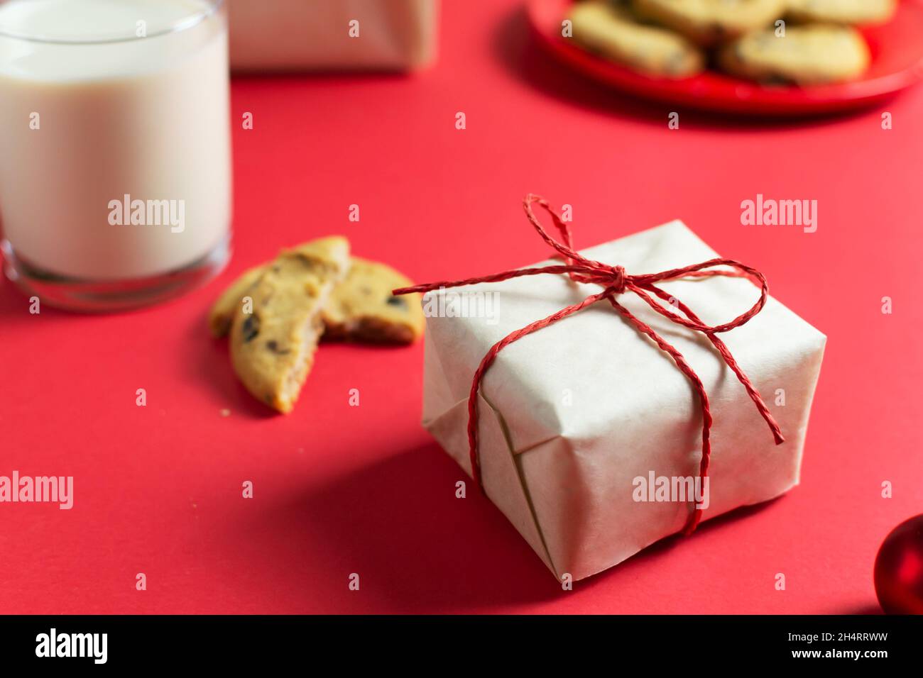 Ein kleines Neujahrsgeschenk ist in Bastelpapier, einem Teller mit Keksen und einem Glas Milch auf rotem Hintergrund verpackt. Horizontale Ausrichtung. Stockfoto