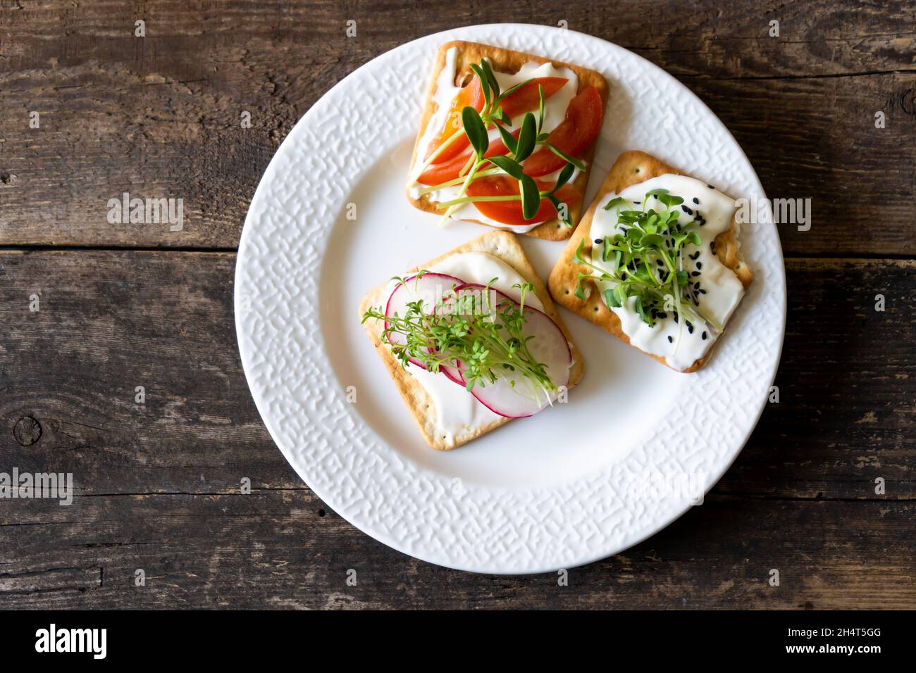 Set Bruschett mit Frischkäse und Tomatenaufstrich. Rettich, Mikrogrün und Leinsamen in Lebensmitteln. Einfaches Essen auf dem Teller. Das Frühstück auf einem weißen Teller war von oben aus zu sehen. Holzhintergrund. Cracker Stockfoto