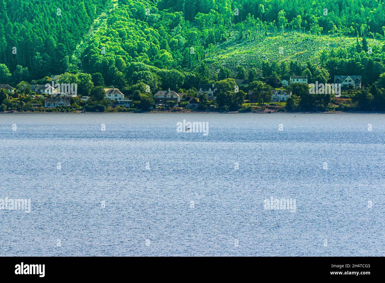Blick über einen See in Schottland. Loch Ness bei Tag mit dem Boot. Häuser am Ufer mit Wald und Bergen im Hintergrund im Sommer. Grüne Bäume auf dem Stockfoto