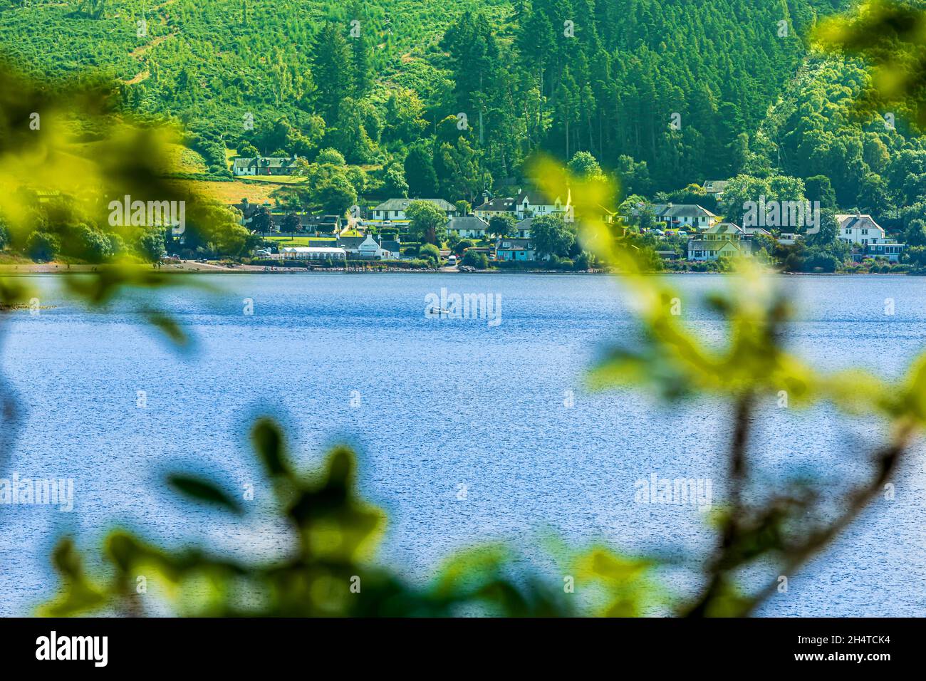 Loch Ness von Schottland im Sommer. Blick durch die Äste des Sees mit dem Boot. Häuser und Bäume im Hintergrund. Berghänge mit verschiedenen Typen Stockfoto