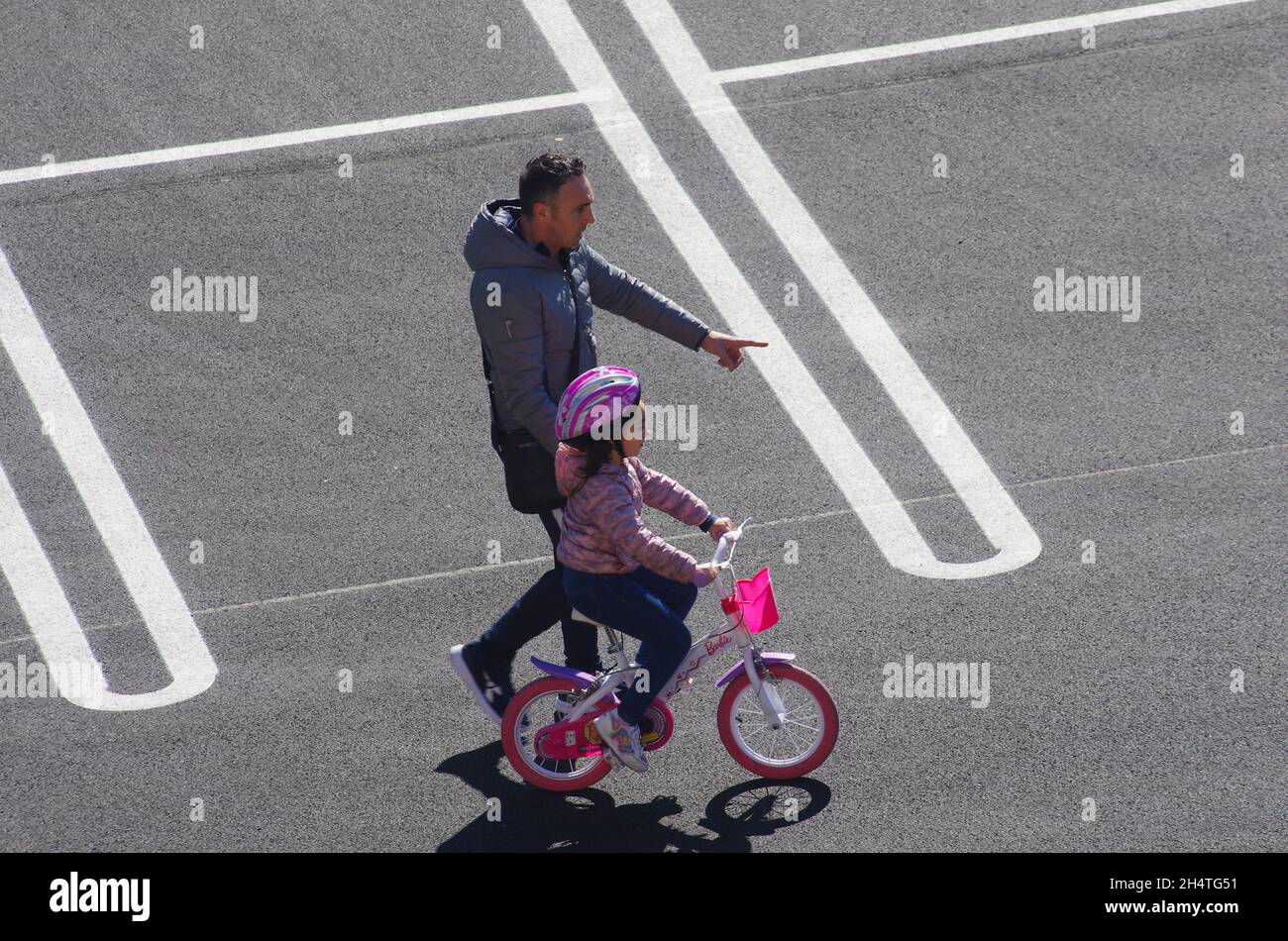 Ein kleines Mädchen mit einem Schutzhelm lernt, mit dem Führer ihres Vaters Fahrrad zu fahren. Stockfoto