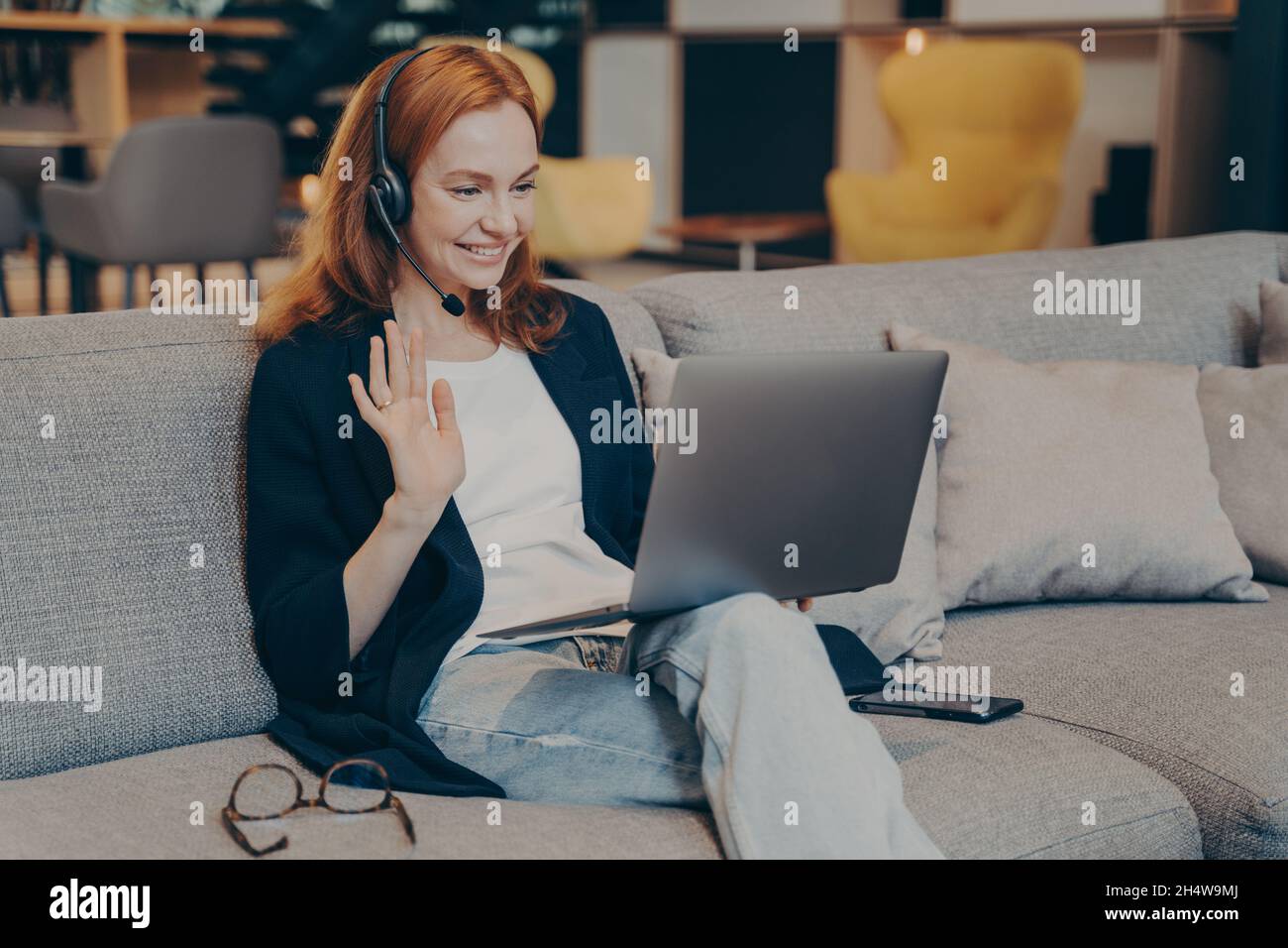 Junge Frau mit Laptop und Headset für Internet-Videoanruf winkt Hand in Grußgeste Stockfoto