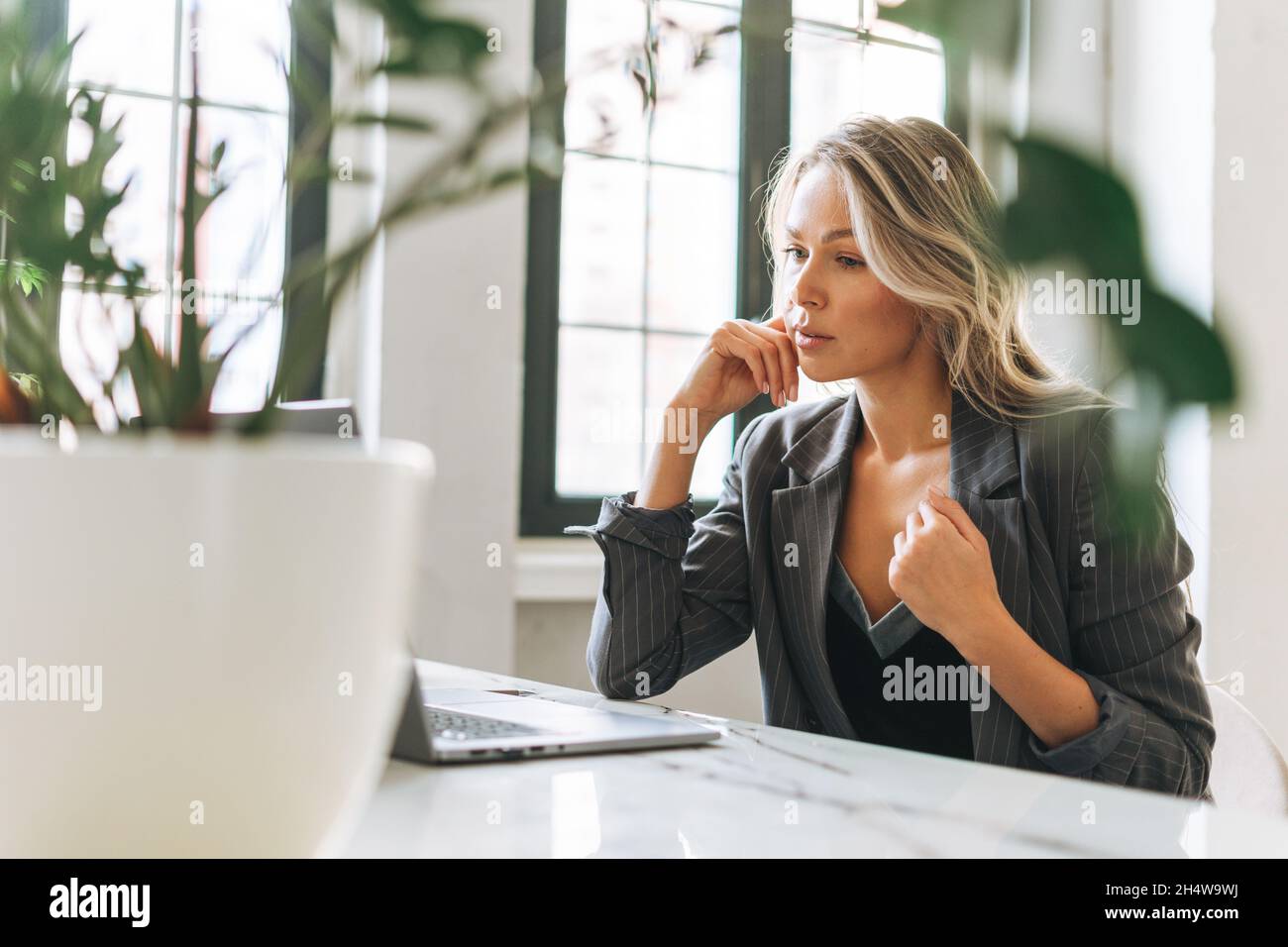 Junge lächelnde blonde Frau mit langen Haaren in stilvollem Anzug, die in einem hellen, modernen Büro am Laptop arbeitet und bei einem Videoanruf spricht Stockfoto
