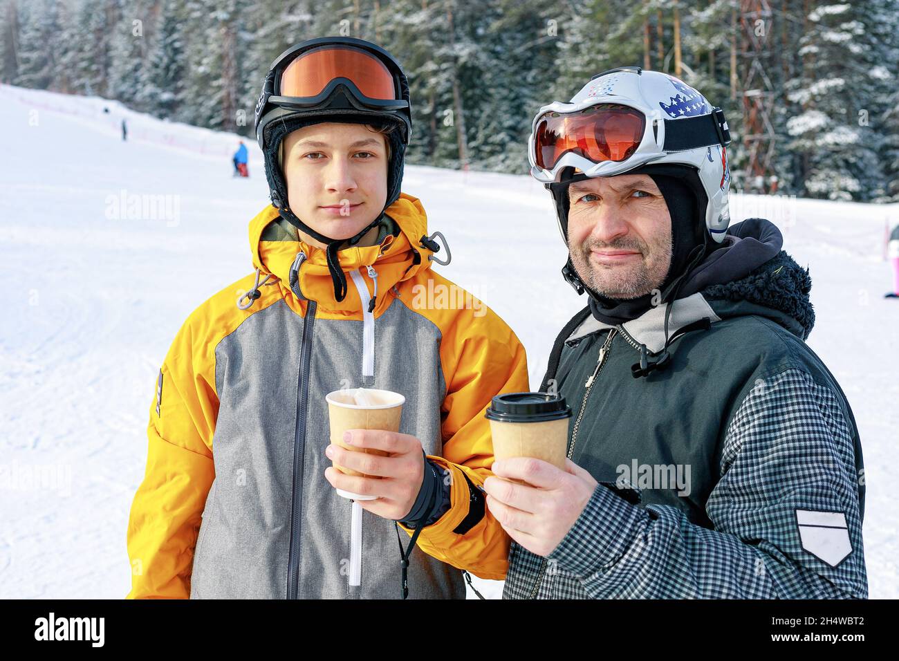 Zwei Snowboarder unterschiedlichen Alters trinken heißen Kaffee. Hintergrund der Skipiste. Vater und Sohn kaukasischen Ethnizität sind im Winter extreme spo beschäftigt Stockfoto