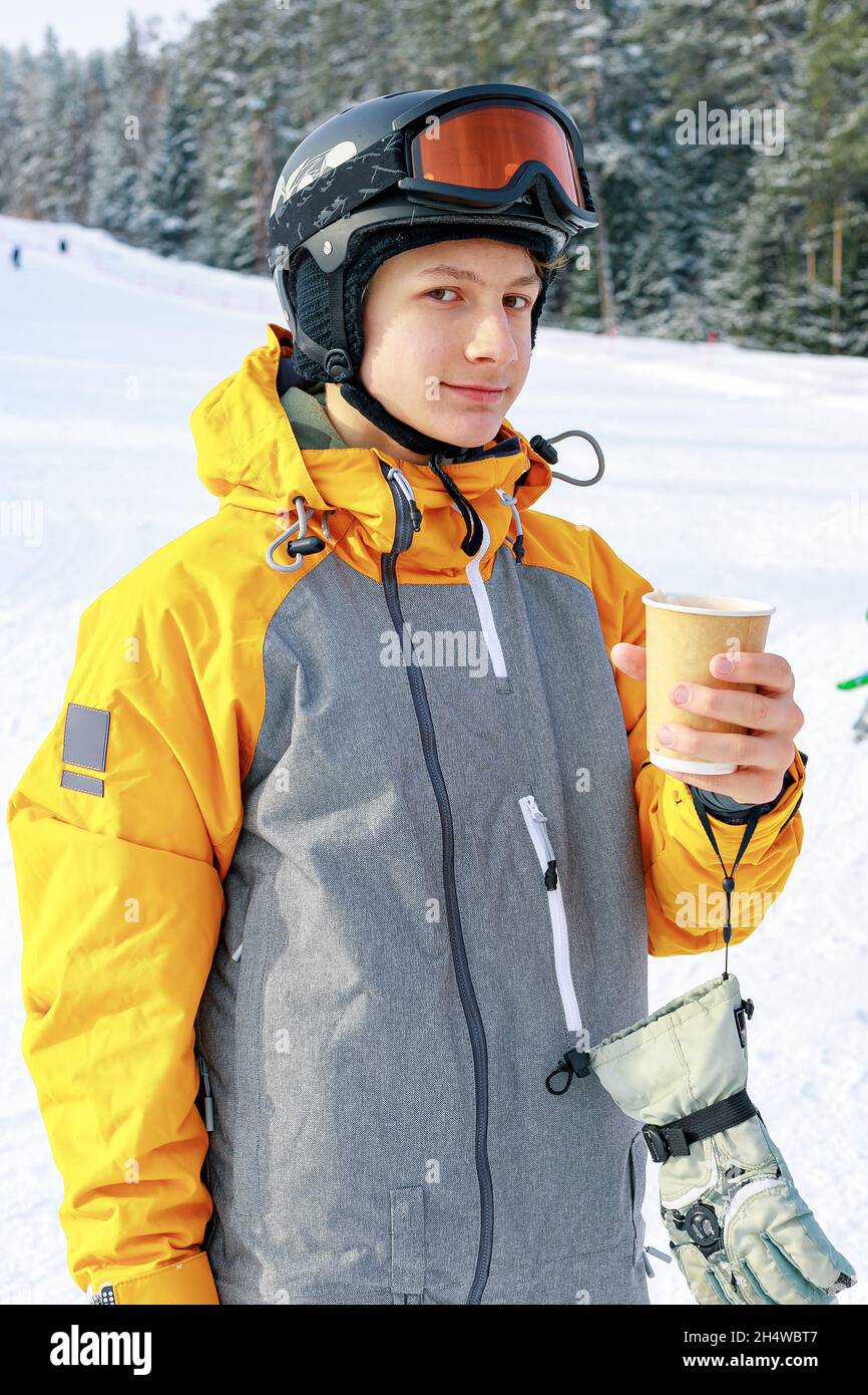 Der Snowboarder trinkt heißen Kaffee. Ein junger Mann aus dem Kaukasus hält einen Papierbecher mit einem heißen Getränk in der Hand. Hintergrund der Skipiste. Porträt eines AT Stockfoto