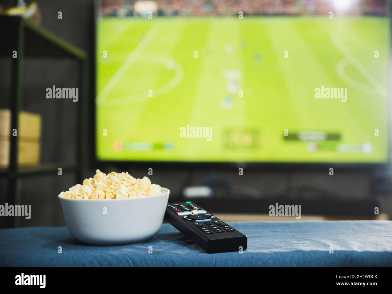 Popcorn- und Fernsehfernbedienung auf dem Hintergrund des Fernsehbildschirms des Fußballprogramms. Fernsehen schauen Entspannungskonzept. Stockfoto