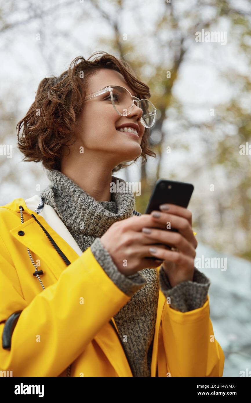 Junges Mädchen in gelbem Regenmantel mit Smartphone in der Hand zu Fuß in der Stadt. Junge schöne braune haarige Frau trägt Regen Outfit während gehen auf stroe Stockfoto