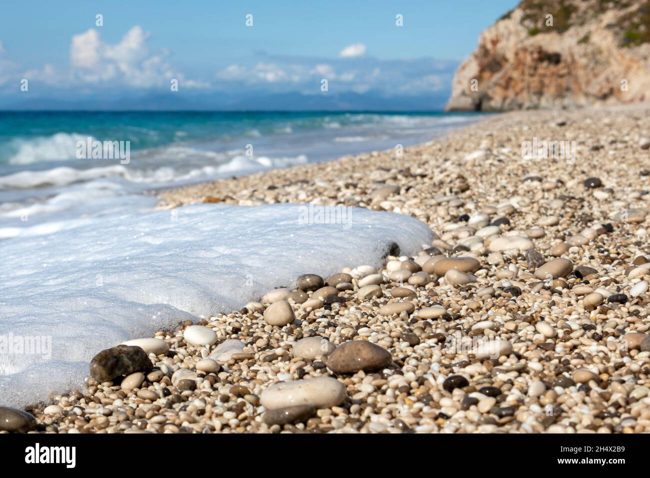 Weißer Schaum auf nassen, glänzenden Kieselsteinen aus nächster Nähe mit azurblauem Meer, stürmischen Wellen, epischen Wolken und felsigen Klippen. Mylos Strand an der Küste der Insel Lefkada in Gr Stockfoto