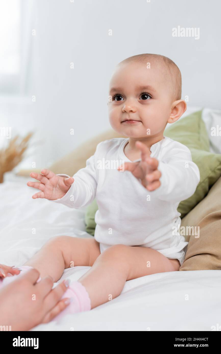 Baby Mädchen sitzt auf dem Bett in der Nähe verschwommener Hände der Mutter Stockfoto