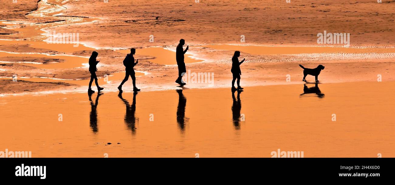 Ein Panoramabild einer Familie von Urlaubern, die am Fistral Beach in Cornwall spazieren gehen und von einem intensiven Sonnenuntergang umgeben sind. Stockfoto