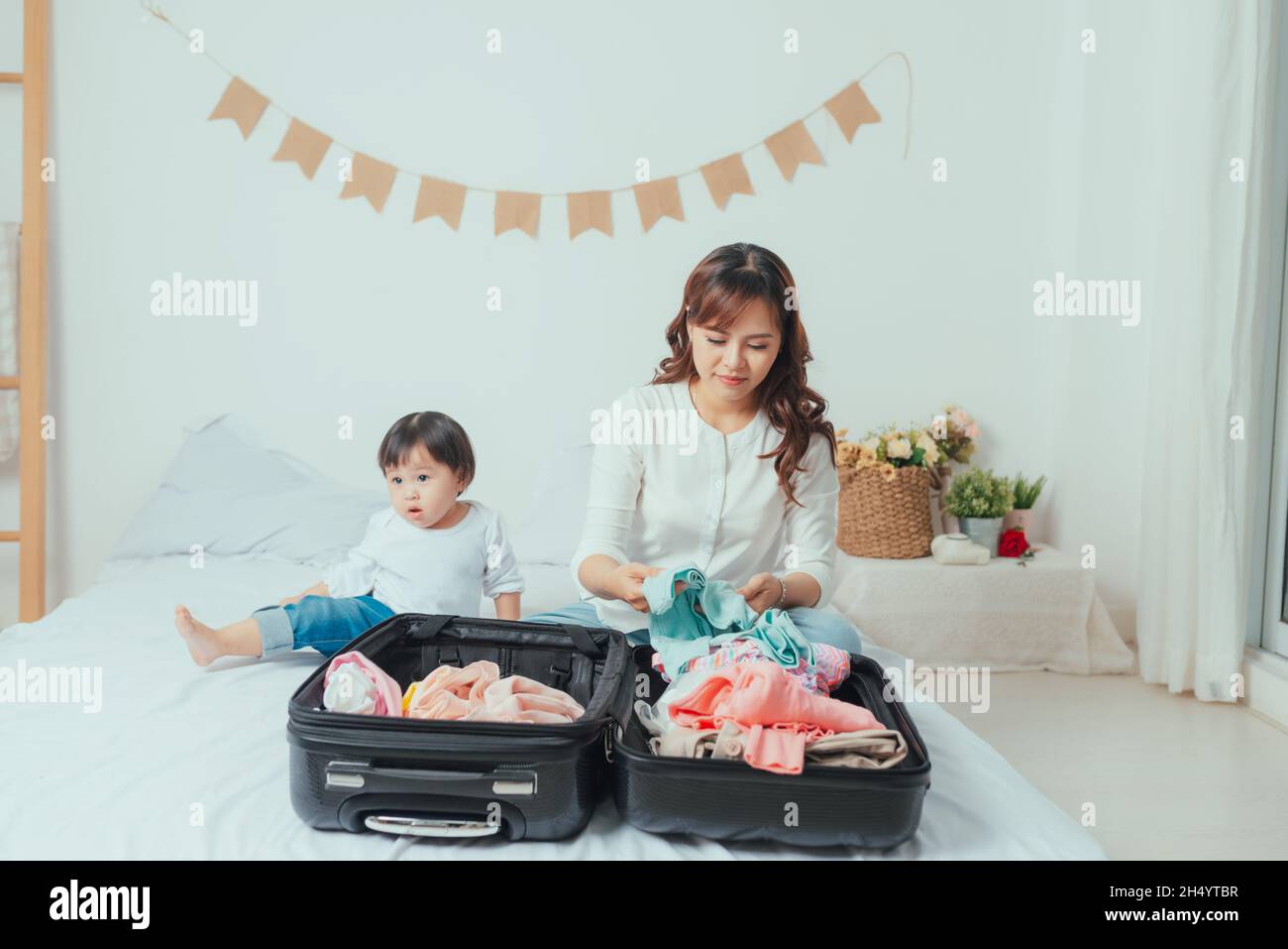 Süßes Baby sitzt neben der Mutter und bereitet Kleidung zum Einlegen in den Koffer vor Stockfoto