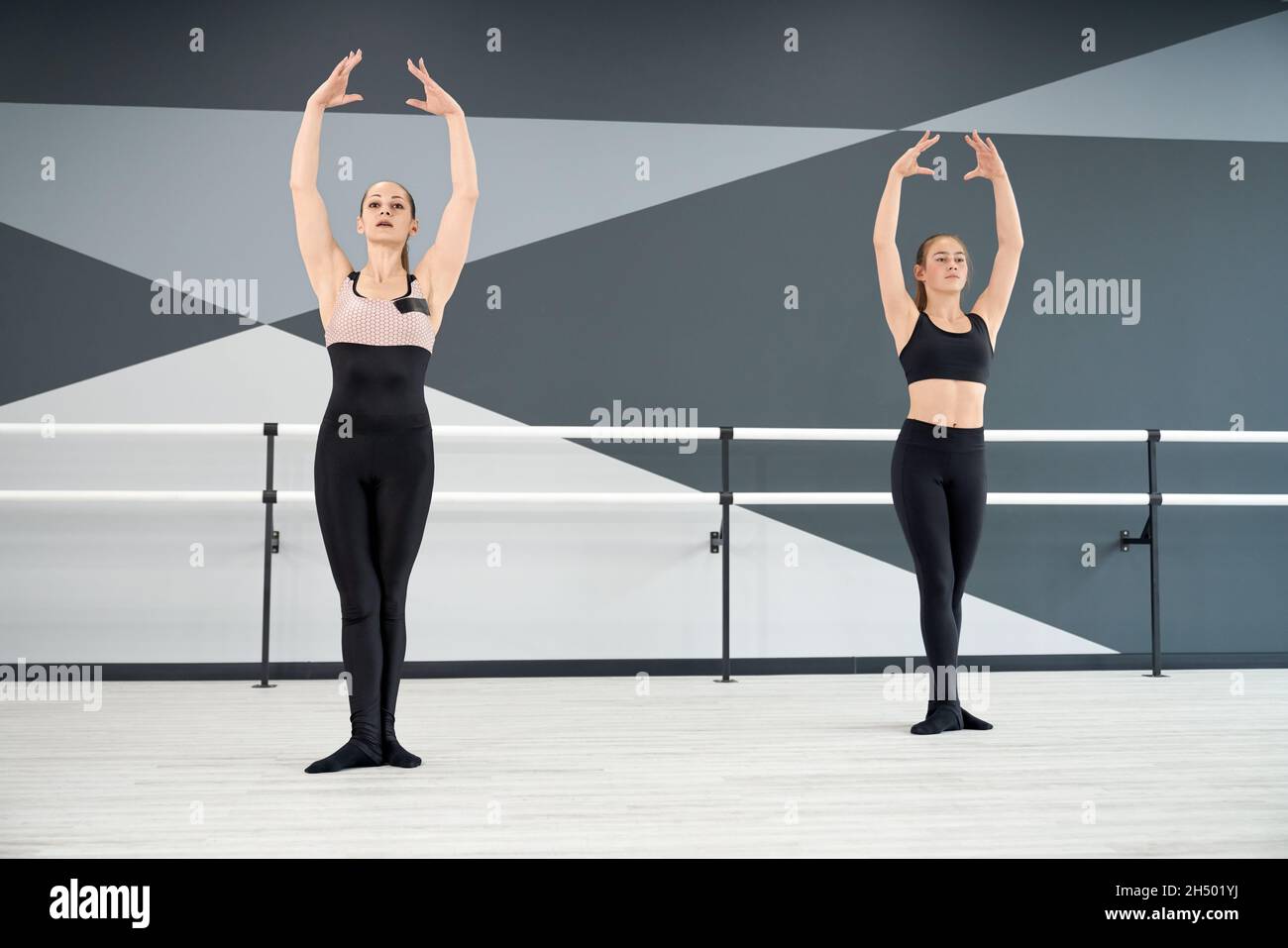 Erwachsene weibliche Ausbilderin hilft jungen hübschen Mädchen in schwarzer Sportkleidung lernen Ballett Pose mit den Händen nach oben. Zwei synchronisierte Frauen üben in der Halle, Hi-Tech-Interieur. Choreographie, Gymnastik-Konzept. Stockfoto