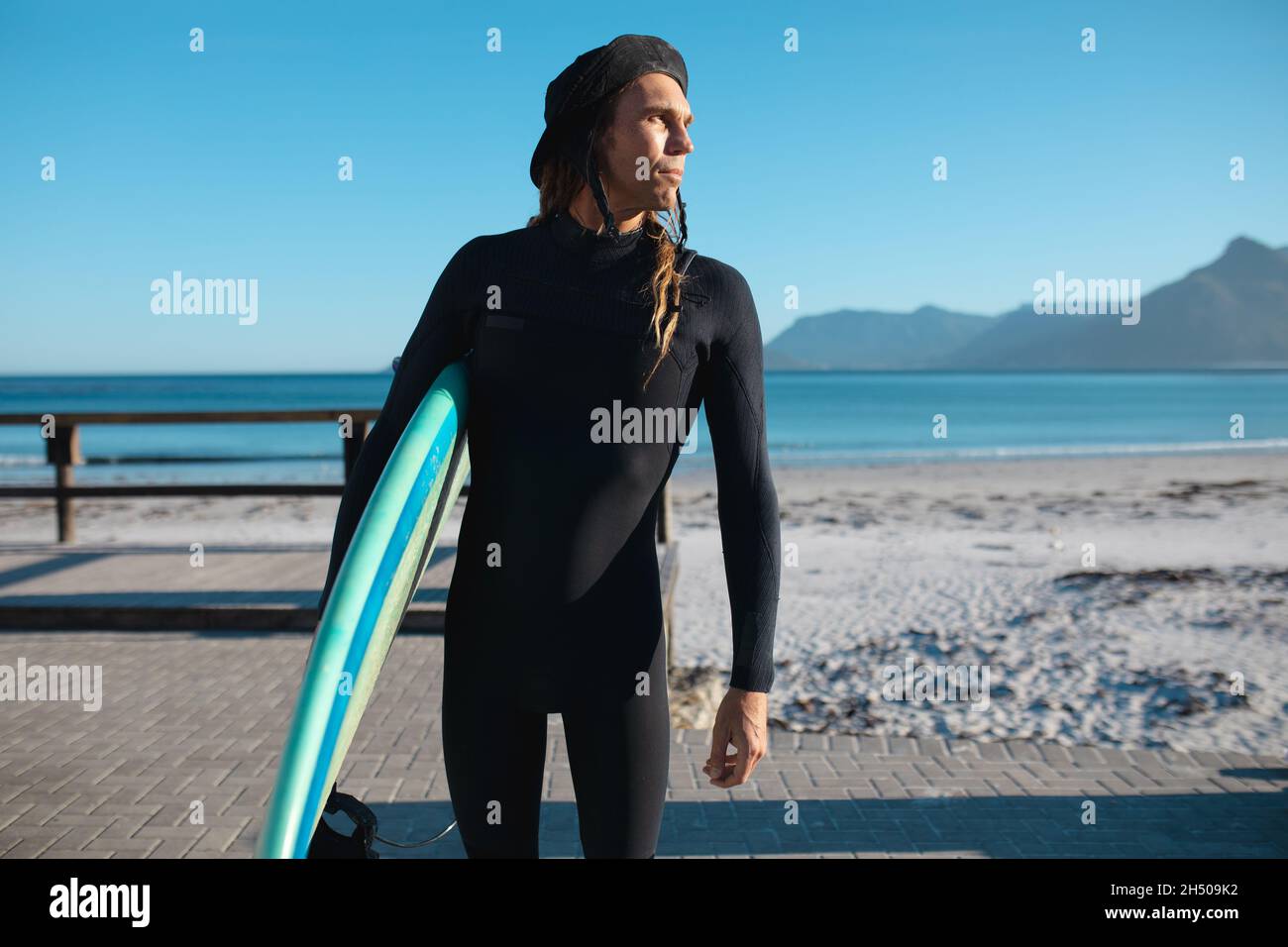 Hipster-Mann schaut weg, während er mit Surfbrett am Strand gegen klaren blauen Himmel steht Stockfoto