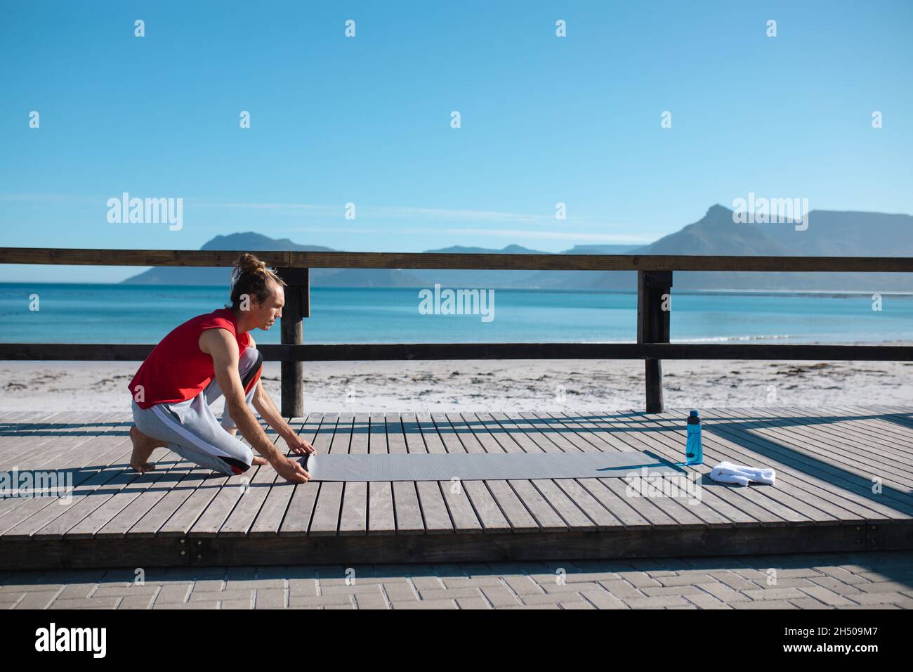 Mann kniet beim Legen einer Yogamatte für das Training am Strand gegen den Himmel mit Kopierraum Stockfoto Mann kniet beim Legen einer Yogamatte für das Training am Strand gegen den Himmel mit Kopierraum Stockfoto