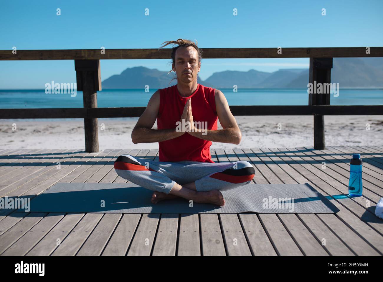 Mann, der Yoga praktiziert, während er mit gekreuzten Beinen sitzt und die Hände zusammenhält und am Strand auf dem Dielenbrett meditiert Stockfoto