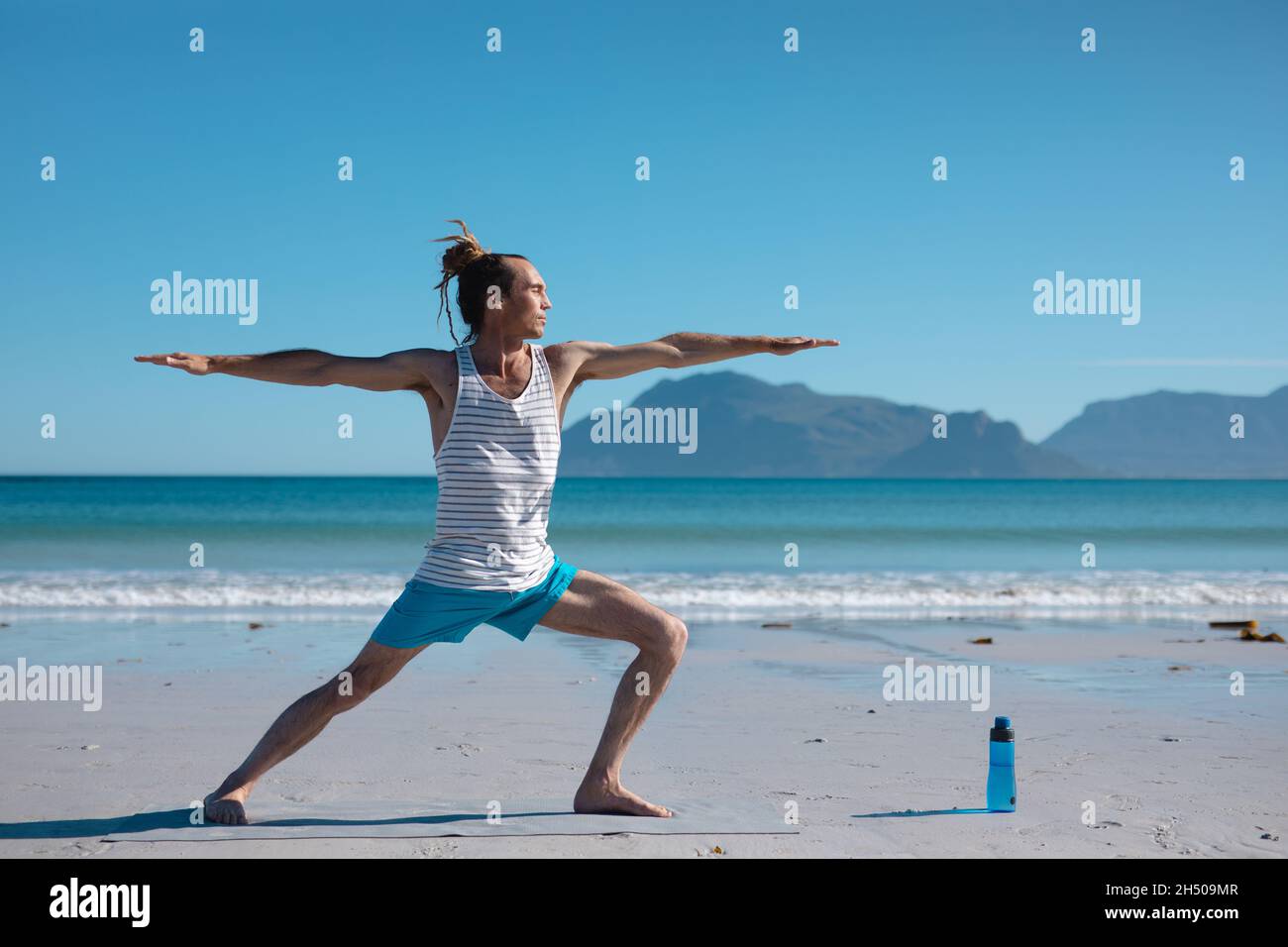 Mann übt Krieger 2 Pose Yoga mit ausgestreckten Armen am Strand gegen Himmel mit Kopieplatz Stockfoto