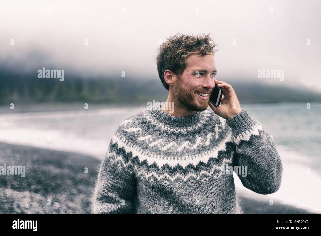 Telefonmann im Winterpullover auf dem Smartphone beim Spaziergang am schwarzen Sandstrand in Island. Isländische Wollkleidung. Technologie Mobiltelefon. Stockfoto