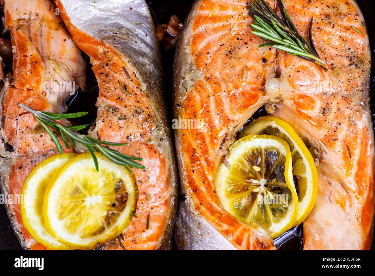 Draufsicht auf frisch geröstete Fischsteaks aus rotem Lachs mit Kräutern, Gewürzen und Zitrone auf schwarzem Metallteller im Hintergrund in der Küche. Stockfoto