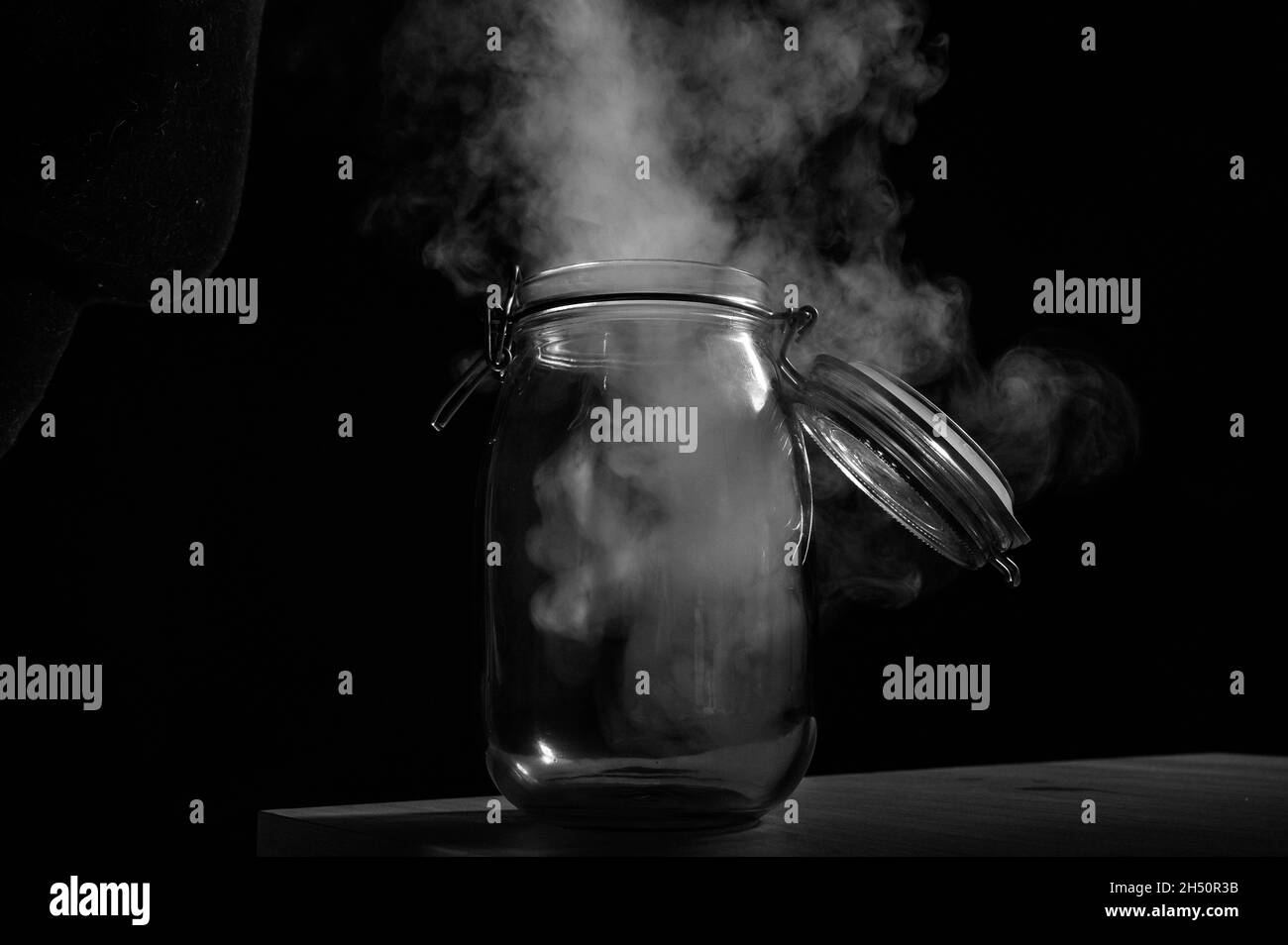 Glas mit einem Deckel, der mit Rauch auf schwarzem Hintergrund gefüllt ist. Nebel in einem transparenten Behälter auf dem Tisch. Stockfoto