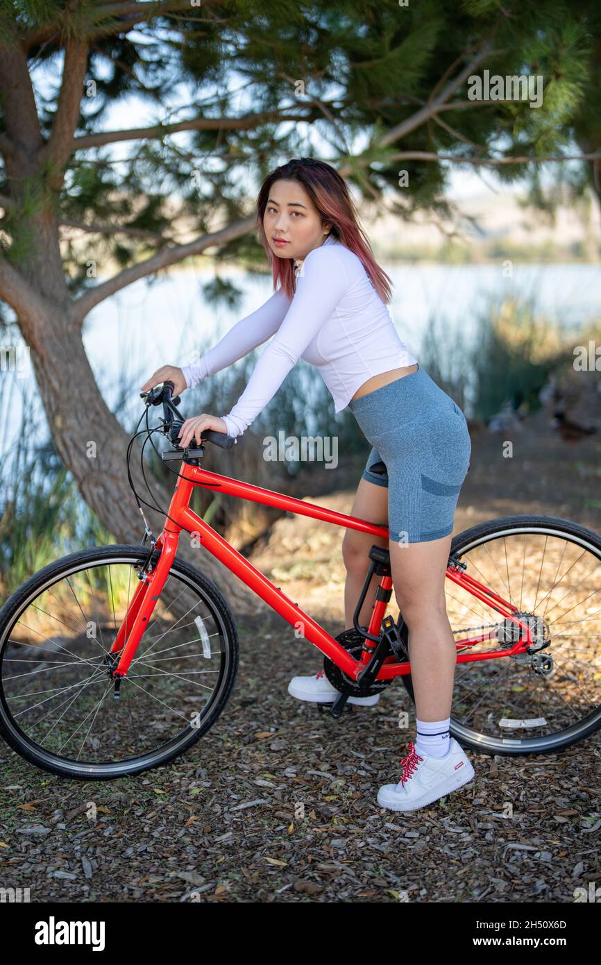 Junge asiatische Frau sitzt auf einem Orange Hybrid Fahrrad vor einem See Stockfoto