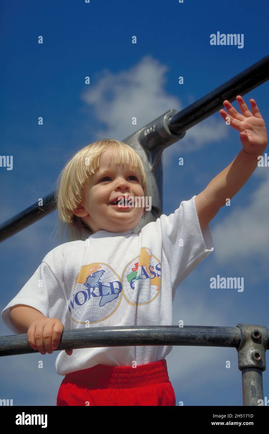 Ein zwei- bis dreijähriges Kleinkind winkt und lächelt von der Oberseite der Spielgeräte gegen einen blauen Himmel. Stockfoto