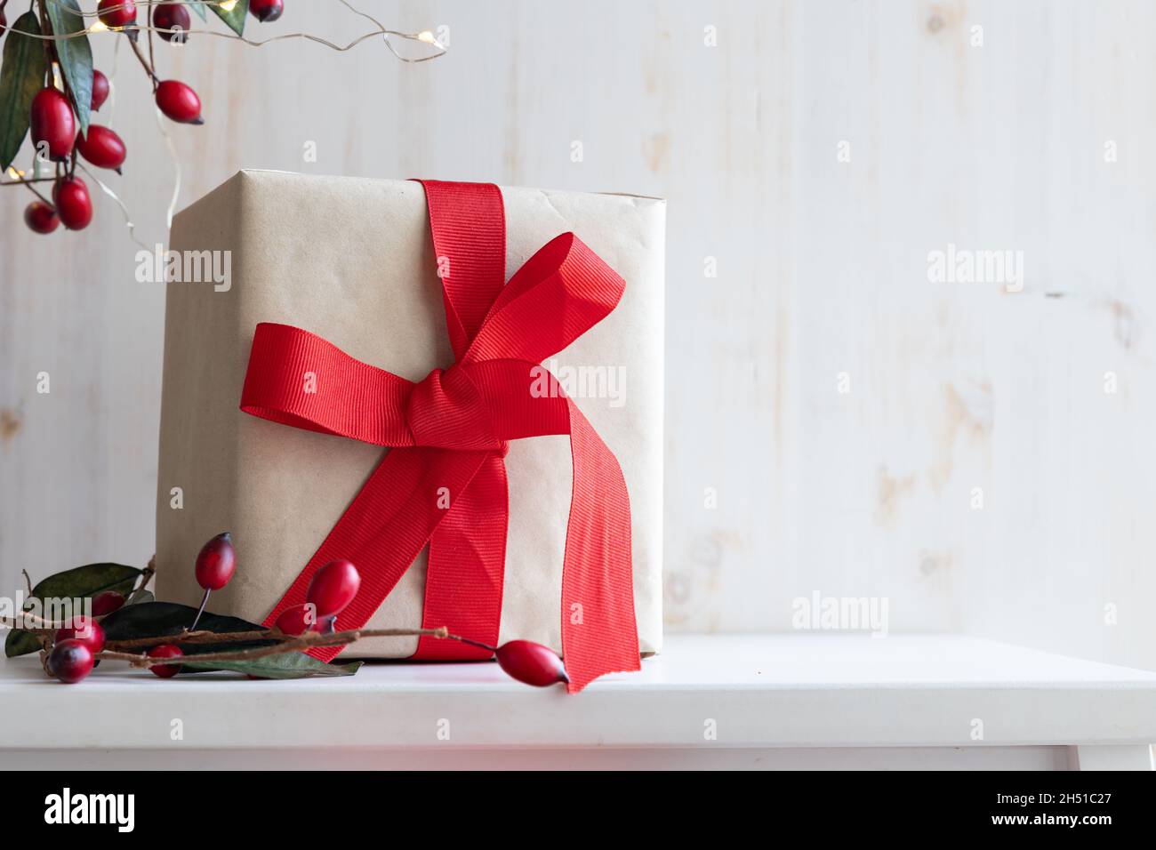 Ein Weihnachtsgeschenk mit braunem Geschenkpapier und roter Schleife mit Zweigen mit roten Beeren und kleinen weißen Lichtern auf Weiß mit Kopierfläche Stockfoto