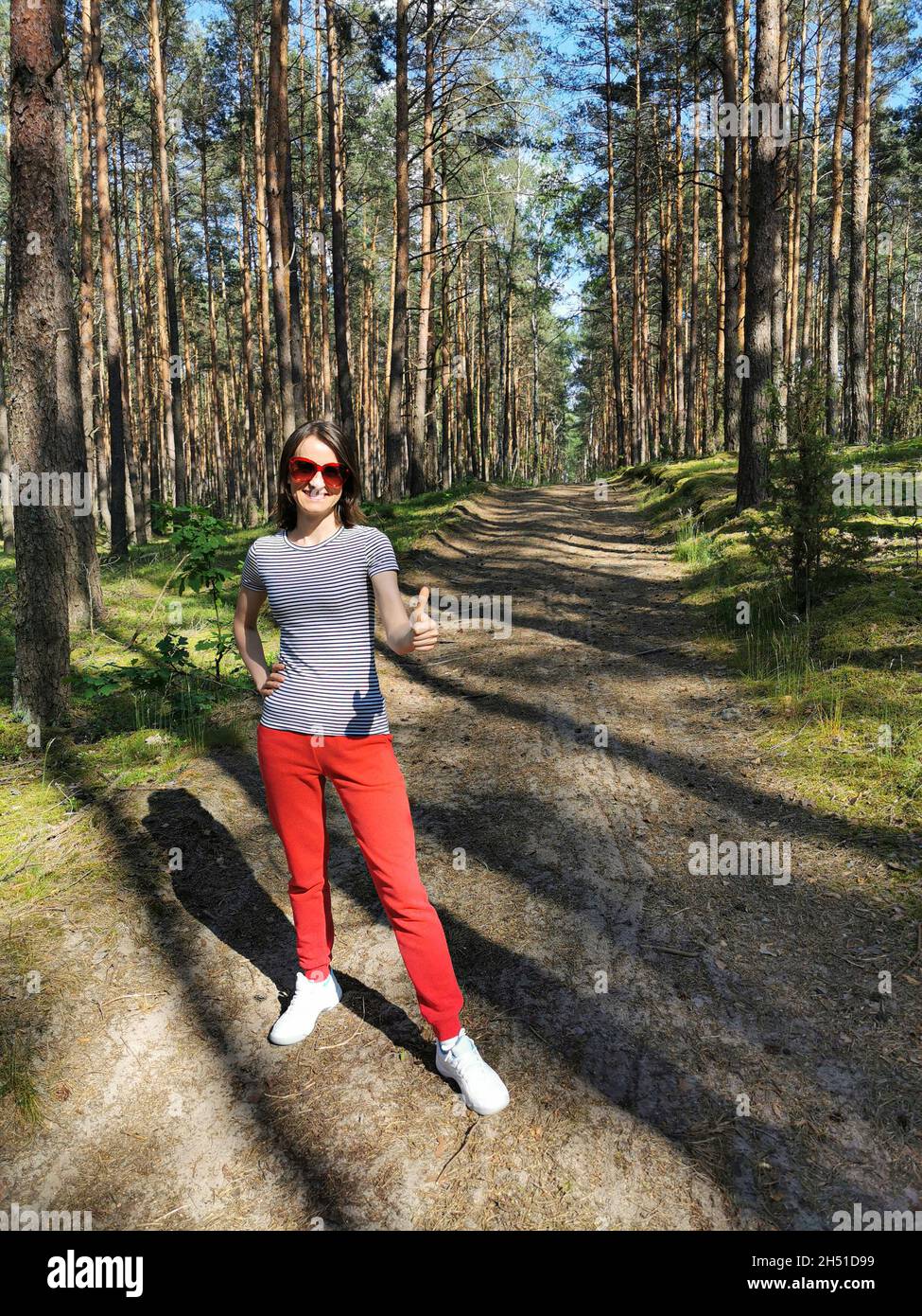 Lächelndes junges Mädchen im Wald zwischen Kiefern mit Daumen nach oben. Schöner sonniger Wald mit hohen Bäumen Stockfoto