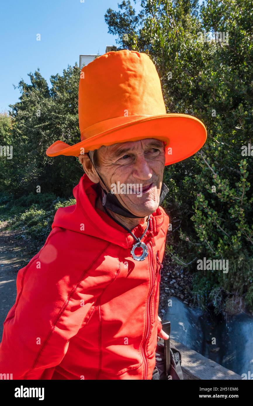 Rand, der Straßenmensch, gekleidet in leuchtend roten und orangen Farben, geht mit seinem sehr hohen orangen Hut entlang der State Street in Santa Barbara County. Stockfoto