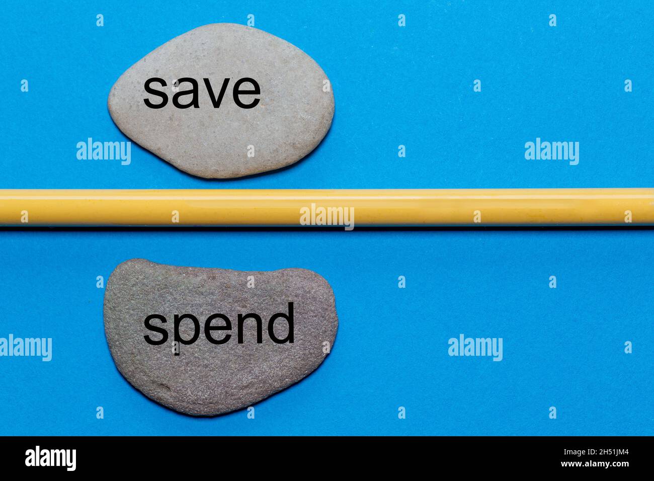 Die Worte Save and Spend sind auf natürlichen glatten Steinen geschrieben, die durch einen gelben Bleistift getrennt sind. Der Hintergrund ist blau isoliert und hat viel Platz Stockfoto
