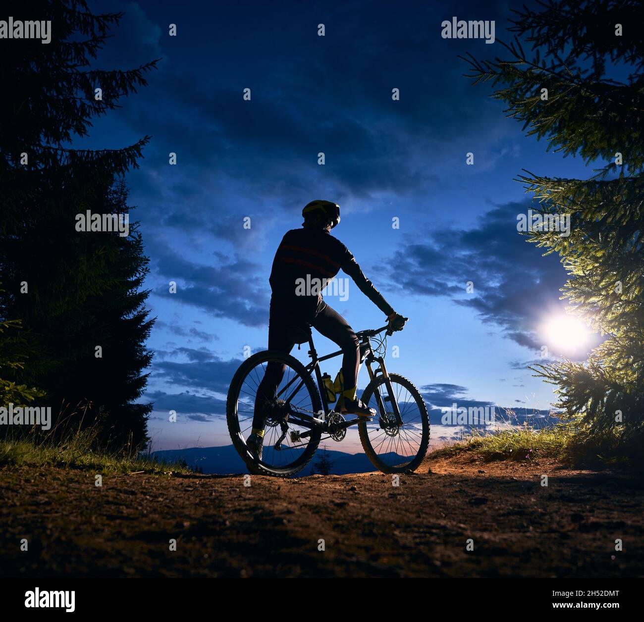 Rückansicht des Mannes, der auf dem Fahrrad unter blauem Abendhimmel mit Wolken sitzt. Silhouette des männlichen Radfahrers Fahrrad auf dem Weg in der Nacht Bergwald. Konzept von Sport, Radfahren und aktiver Freizeit. Stockfoto