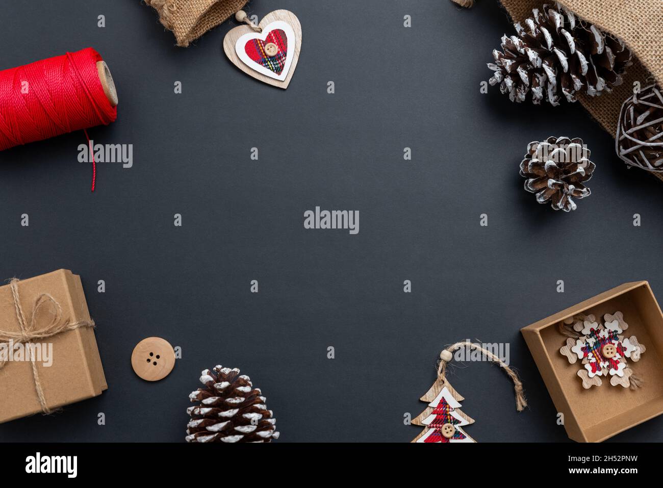 Weihnachtskomposition auf schwarzer Oberfläche mit roten Details auf Weihnachtsdekorationen. Weihnachtsgeschenk, Tannenzapfen, Holzdekorationen, roter Faden, Tailorin Stockfoto