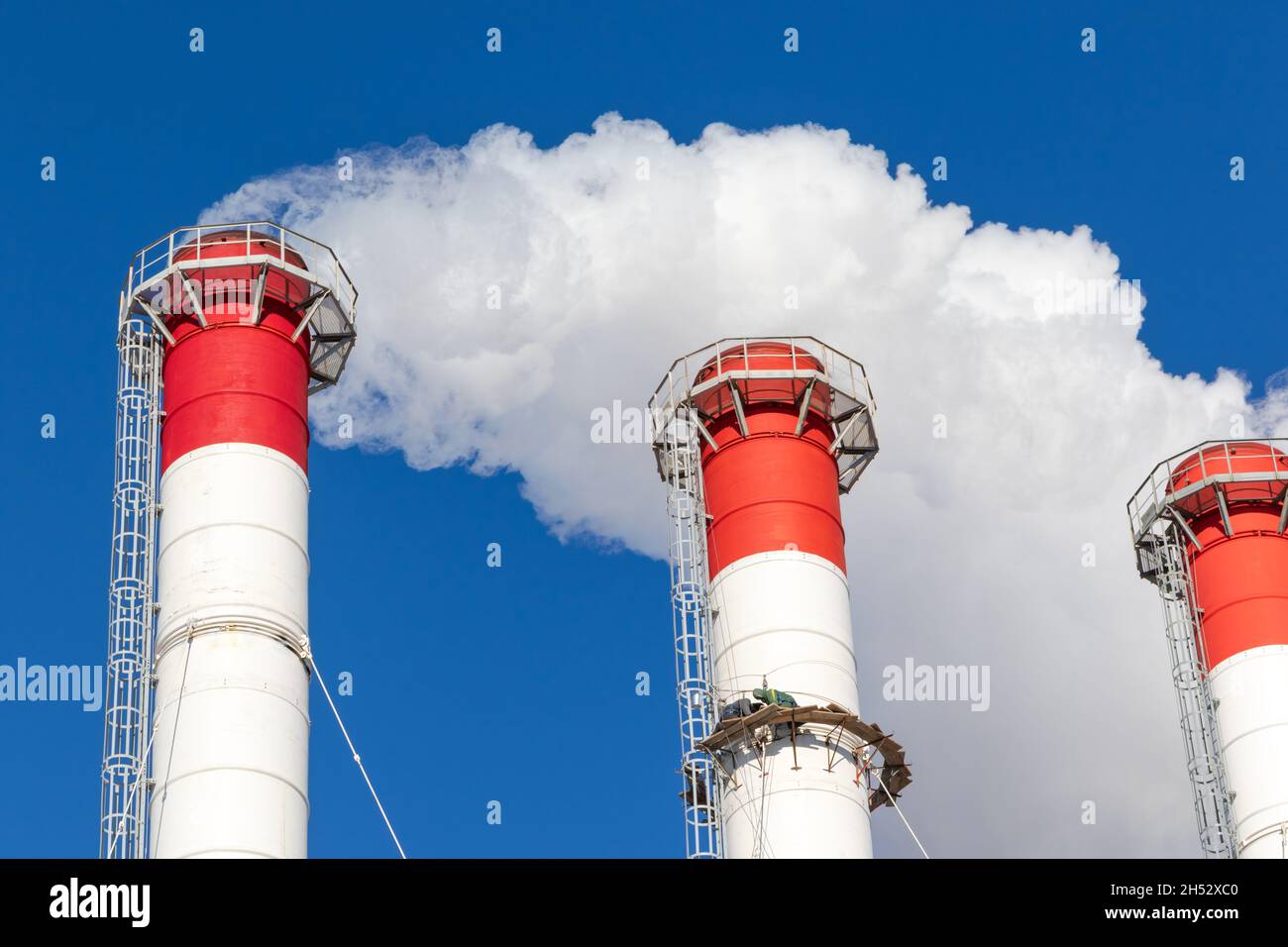 Rot-weiße Schornsteine des Kesselraums, ausgestattet mit einer Ampel. Industriekletterer führen Routinereparaturen durch. Weißer Rauch auf einem blauen Himmel-Rückgro Stockfoto