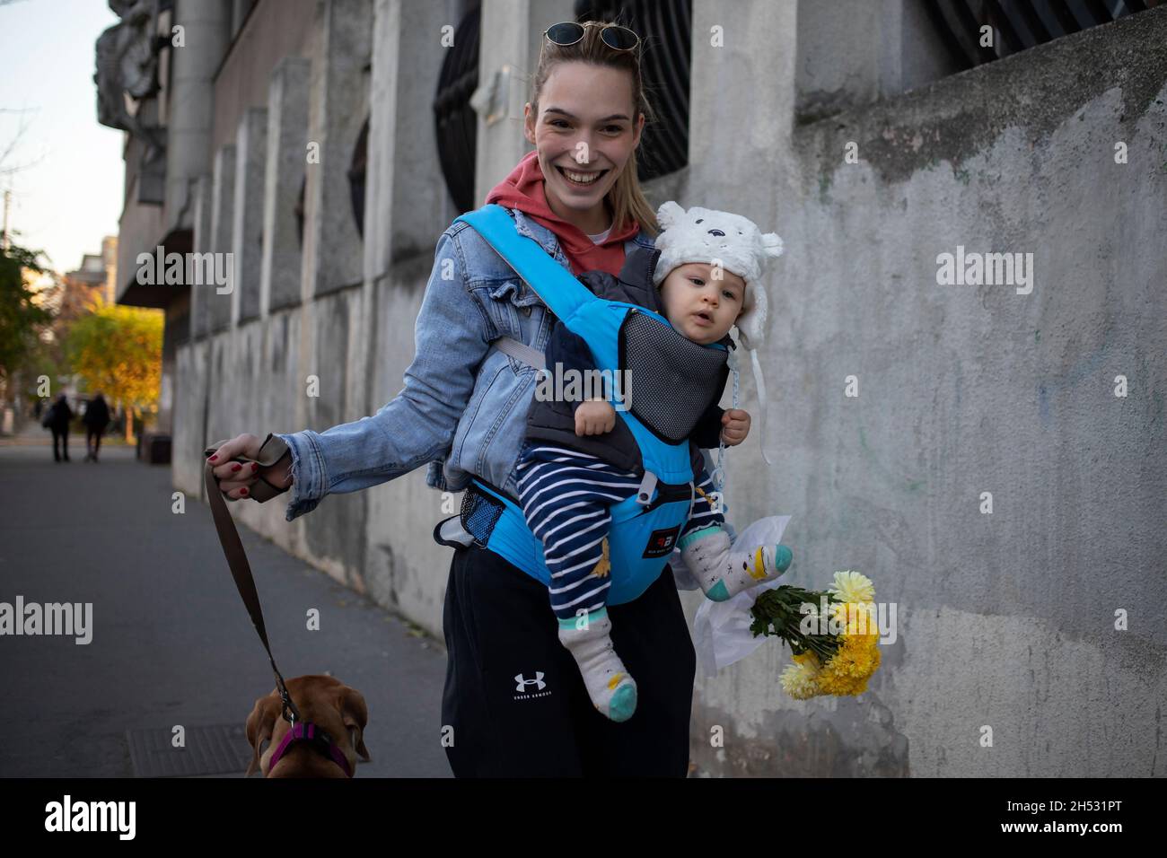 Belgrad, 31. Oktober 2021: Eine junge Frau mit einem Baby in einem Träger und einem breiten Lächeln geht mit einem Strauß gelber Blumen den Hund die Straße entlang Stockfoto