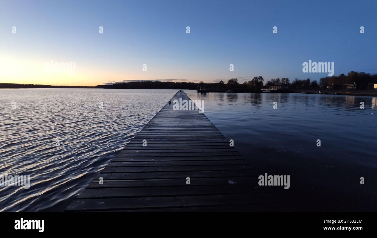 Feuchter hölzerner Pier über der Wolga, der in dunkelblauer Farbe mit dem Wasser zum Horizont zeigt, der durch die untergehende Sonne am Abend erhellt wird Stockfoto