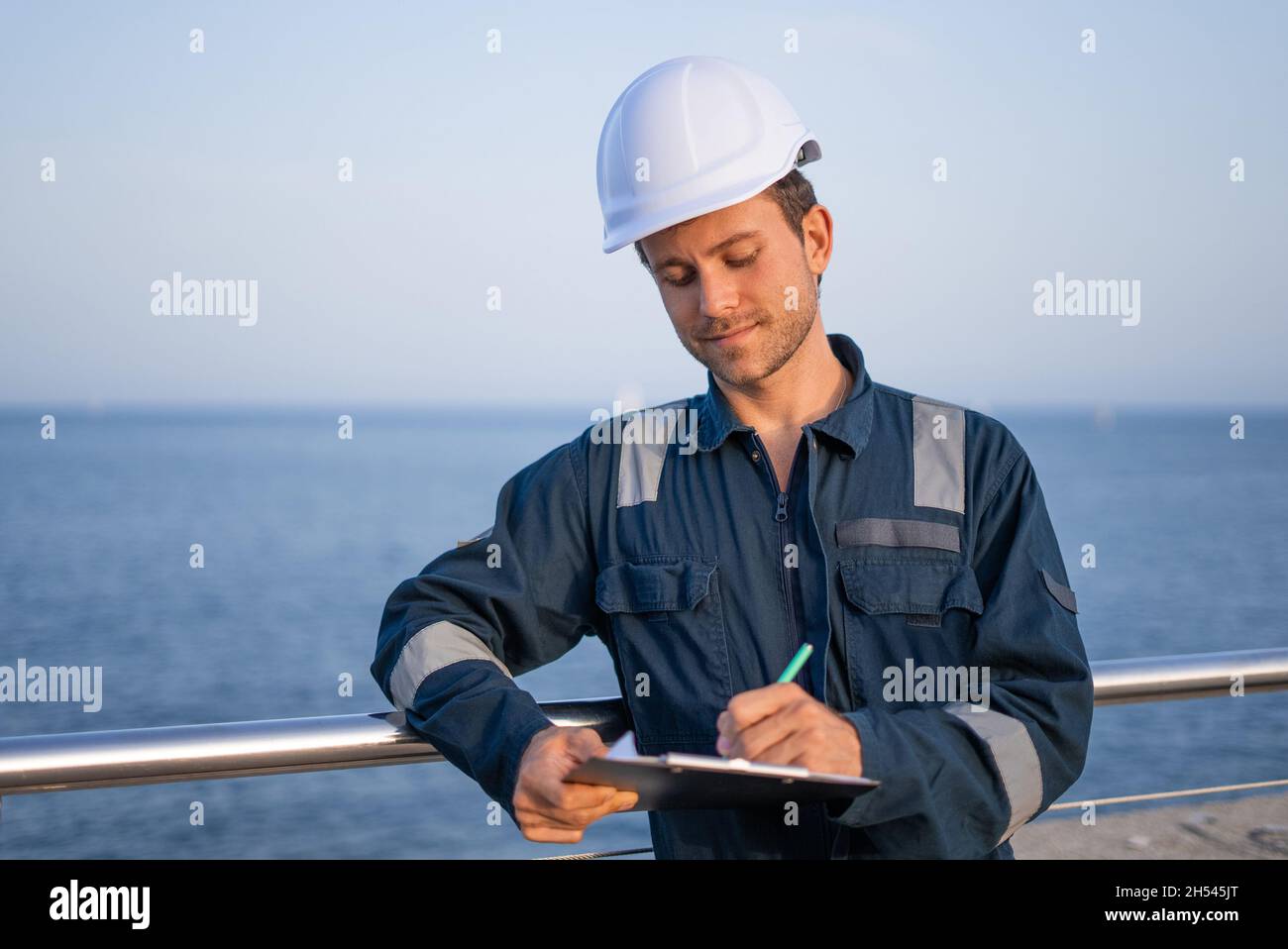 Junge männliche Logistikarbeiter in Schutzhelm und Arbeitskleidung mit Klemmbrett Unterschrift Papiere während der Arbeit im Seehafen Stockfoto