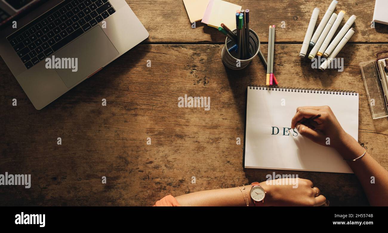 Die junge Frau zeichnet das Wort „DESIGN“, während sie an ihrem kreativen Arbeitsplatz sitzt. High-Angle-Ansicht einer Grafikdesignerin, die an einem neuen Projekt arbeitet Stockfoto