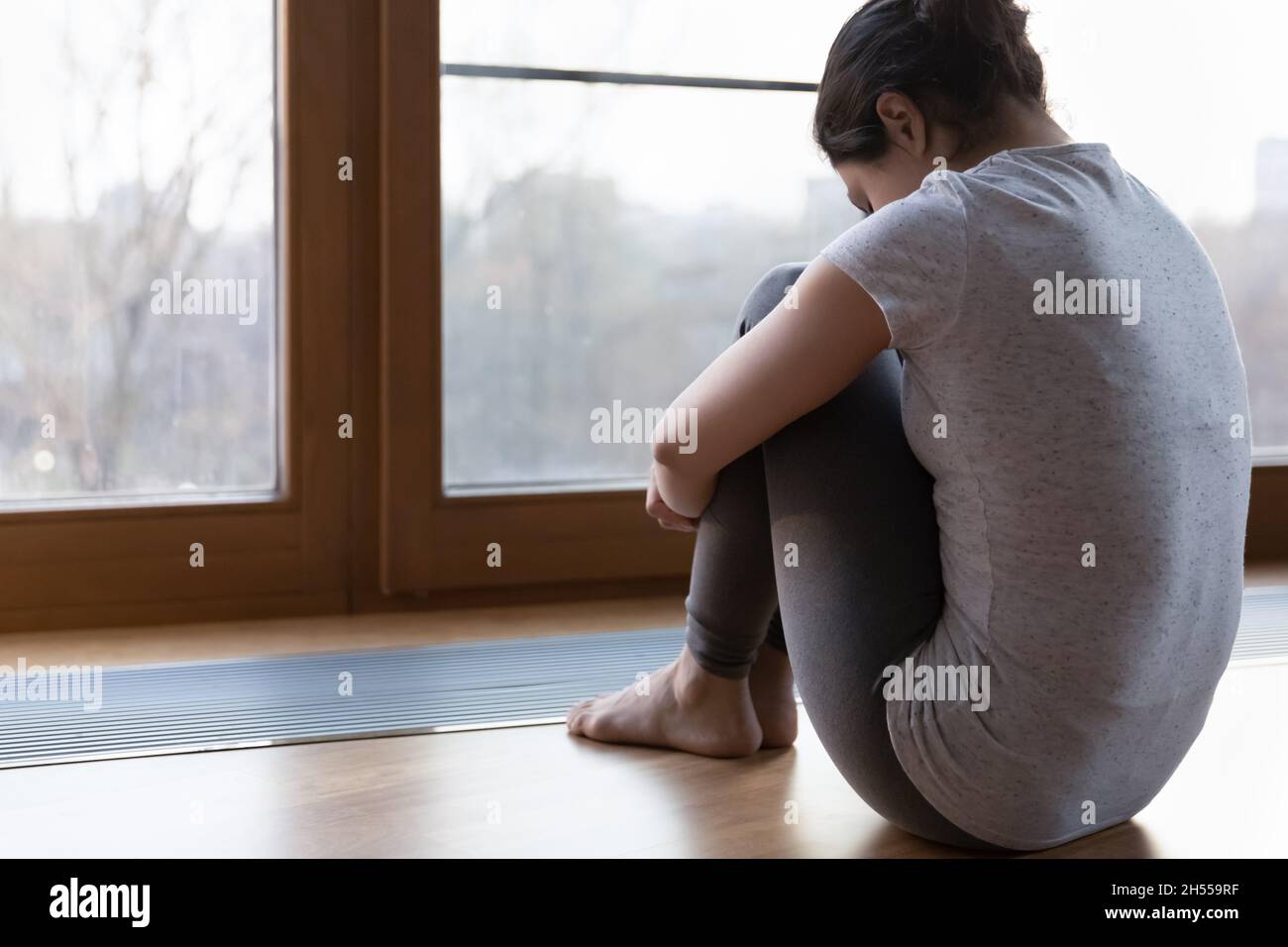 Rückansicht junge unglückliche Frau emb Knie sitzen in der Nähe des Fensters Stockfoto
